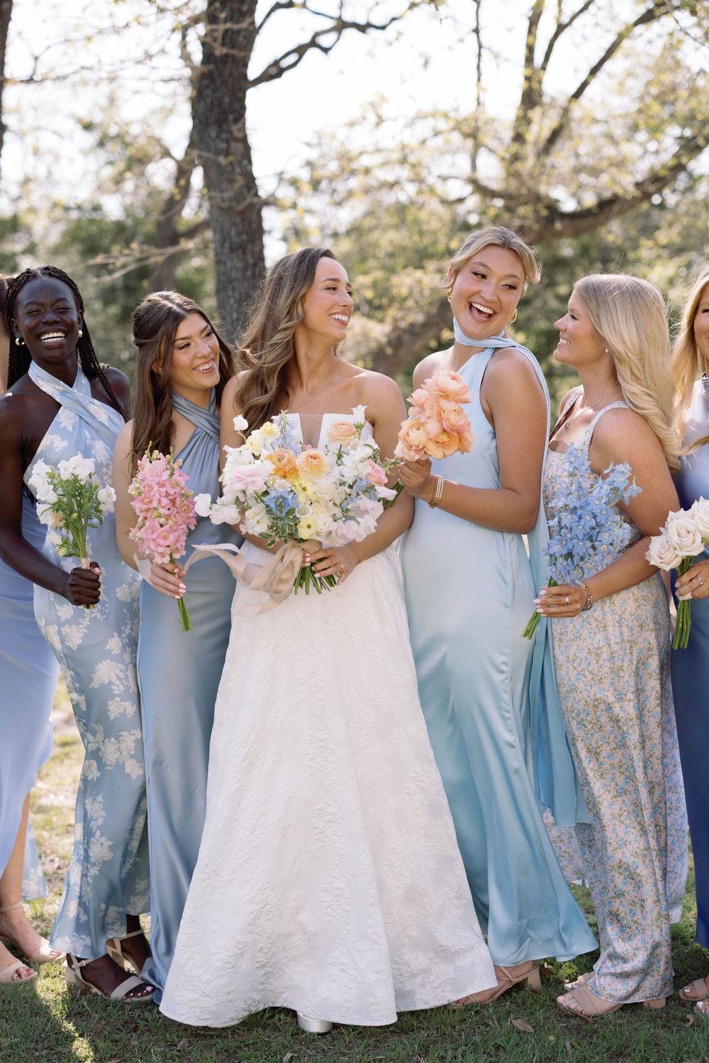 A group of bridesmaids wearing light blue and floral dresses laugh together while holding bouquets during an outdoor ceremony.