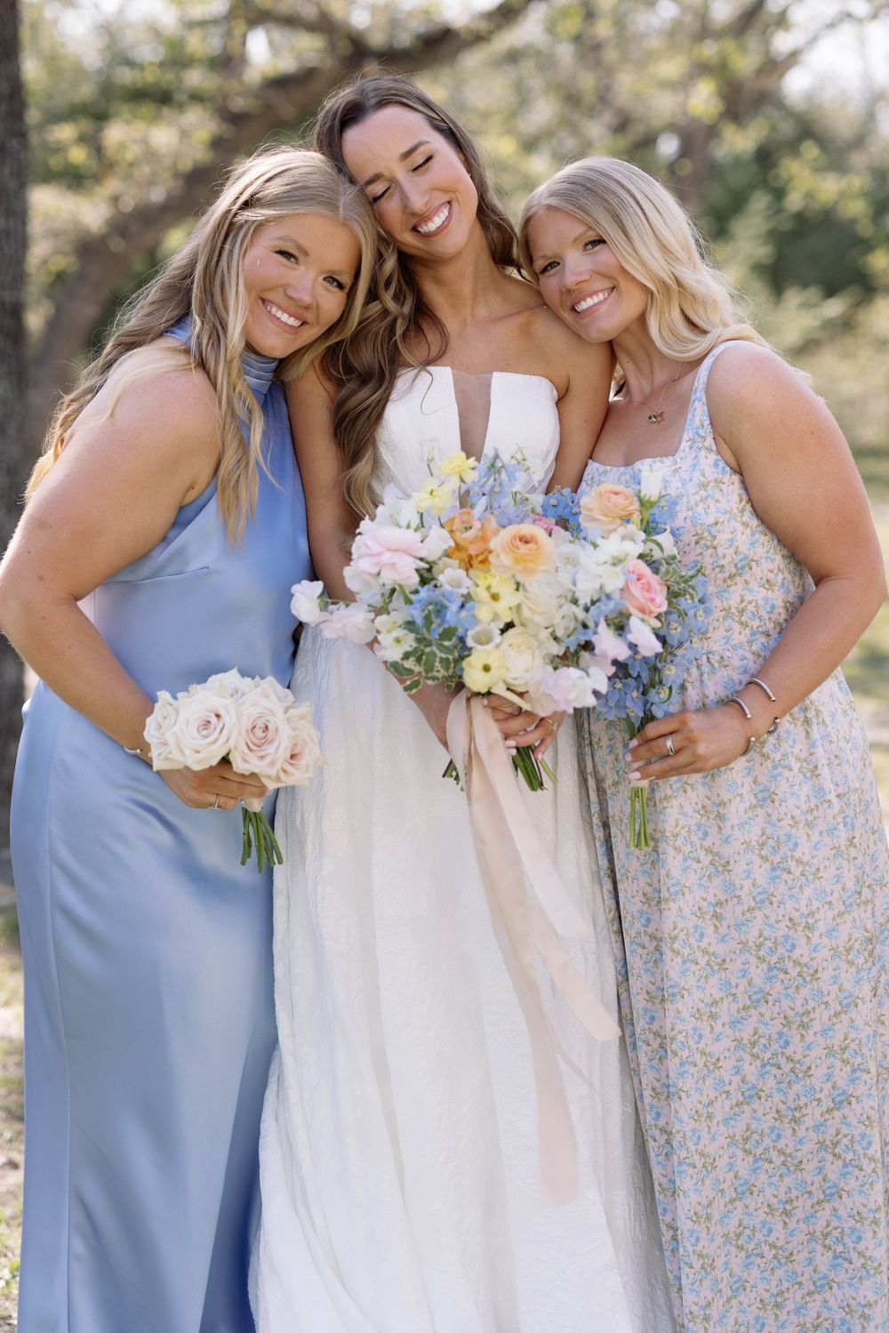 Three bridesmaids pose together at outdoor wedding with blue and floral dresses holding pastel bouquets.