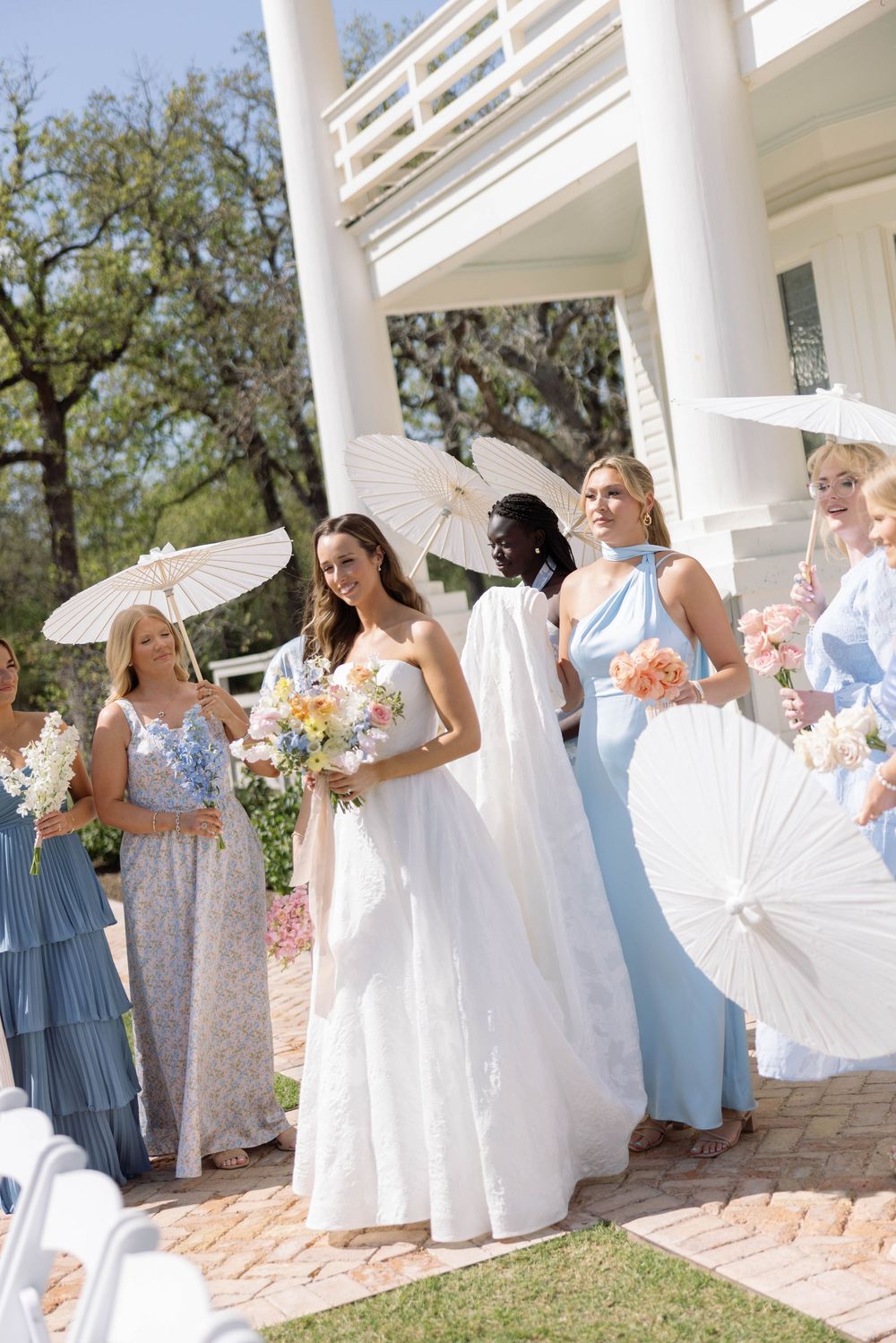 A wedding party stands together on a brick patio lined with white columns wearing light blue and white dresses.