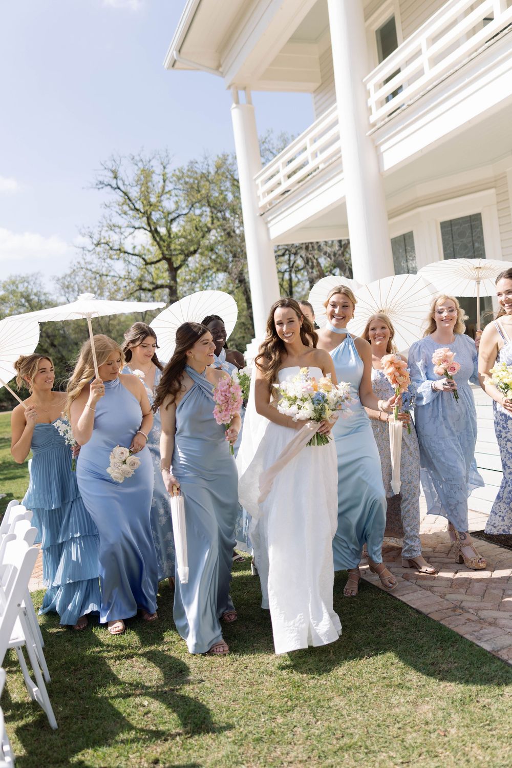Wedding party wearing dusty blue bridesmaid dresses stands together on lawn in front of white columned building.