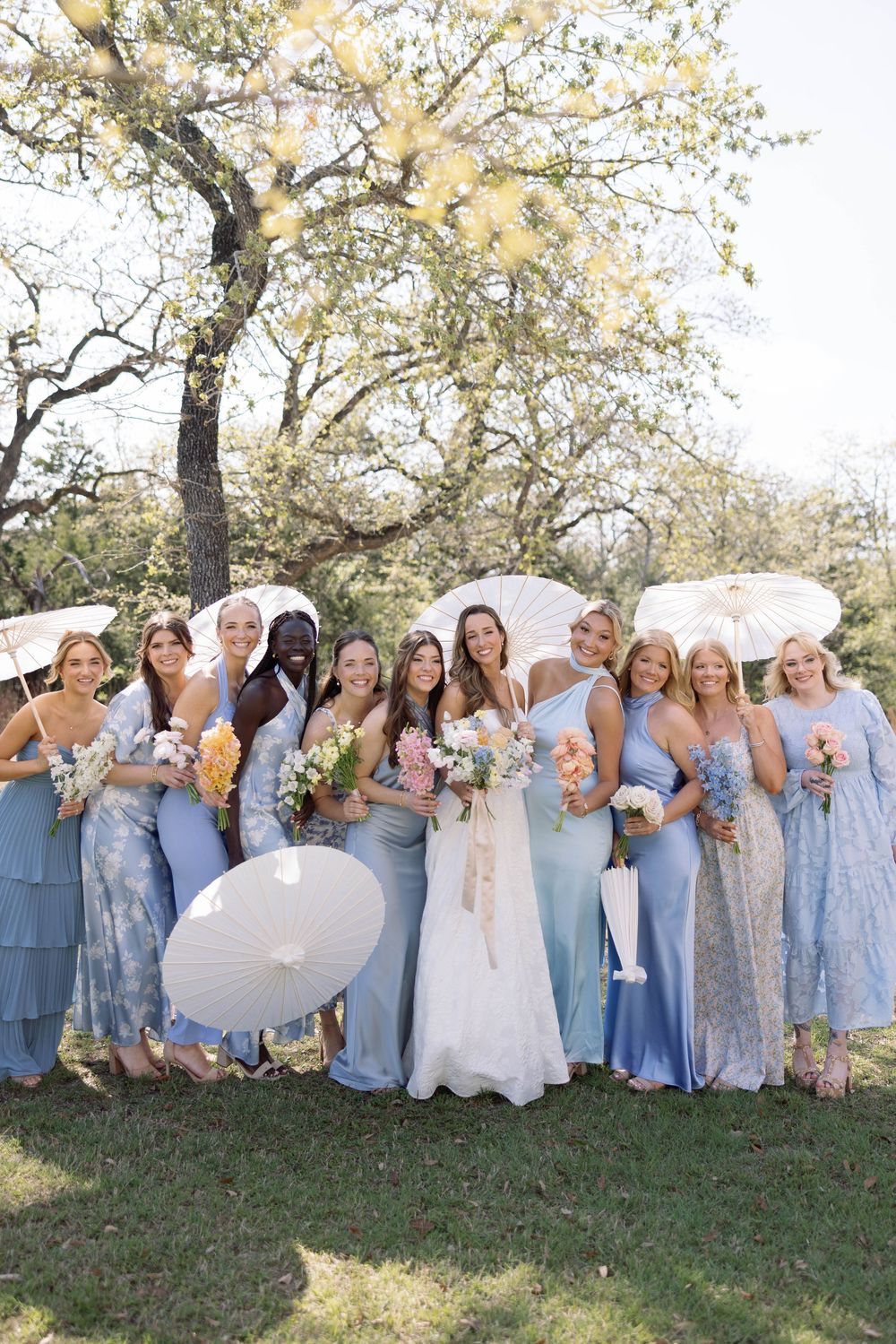 Bridal party in light blue and lace dresses pose with white parasols under blooming spring trees.