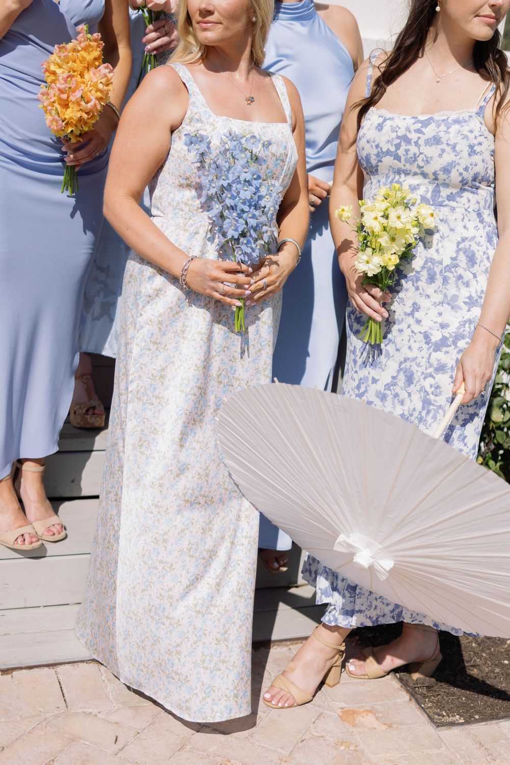Bridesmaids in light blue floral dresses hold colorful bouquets while standing together at an outdoor wedding ceremony.