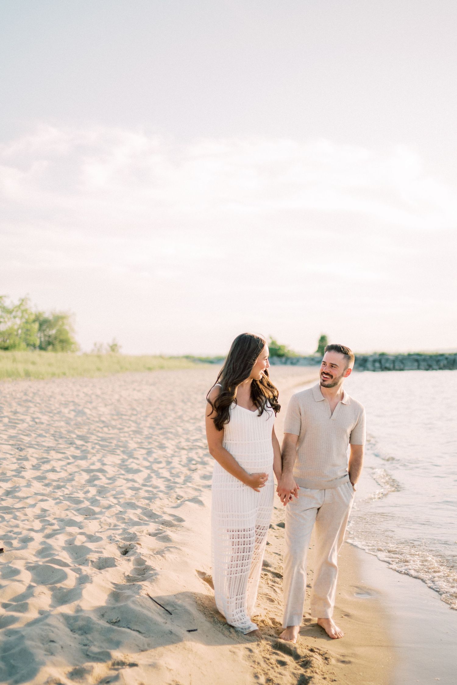 A couple walks hand in hand along a sandy beach at sunset wearing white and beige coordinated outfits.