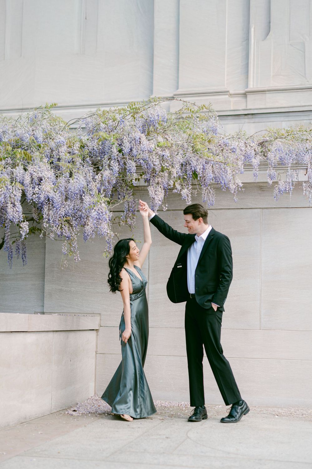 Dancing couple perform playful moves under blooming wisteria against white stone wall.
