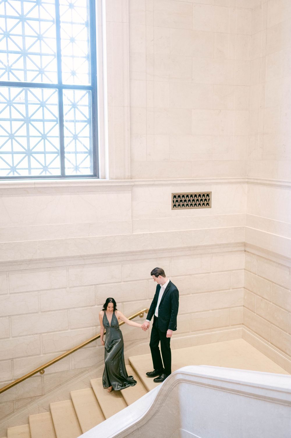 Couple dancing on white marble staircase near geometric window.
