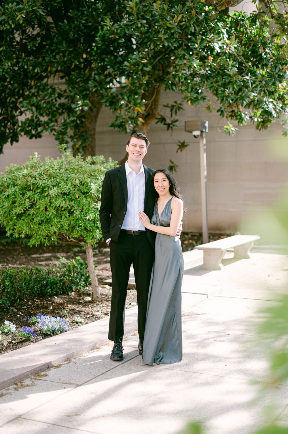 A couple in formal wear posing in a garden setting with blooming flowers.