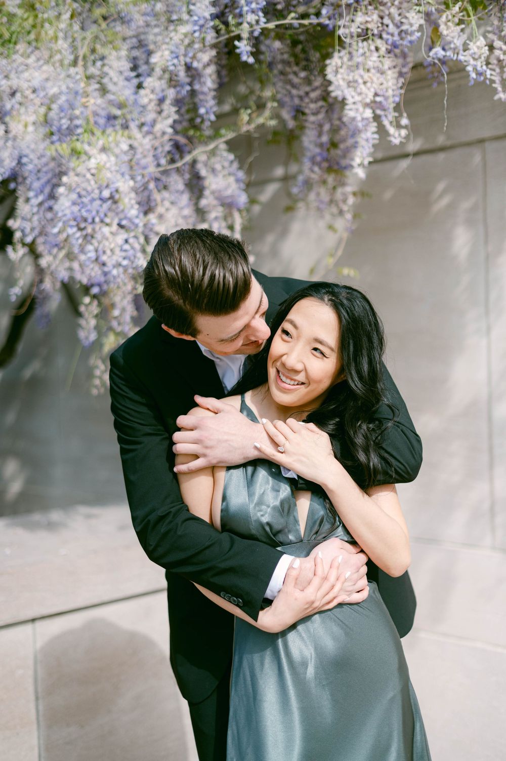 A tender moment shared under blooming wisteria vines against a white wall.