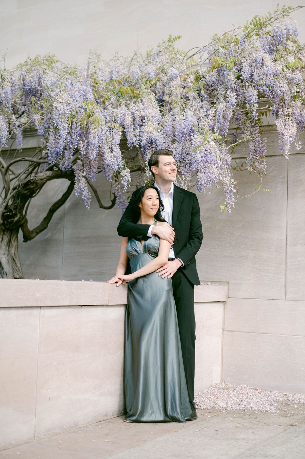 A romantic embrace under cascading purple wisteria flowers against a stone wall.