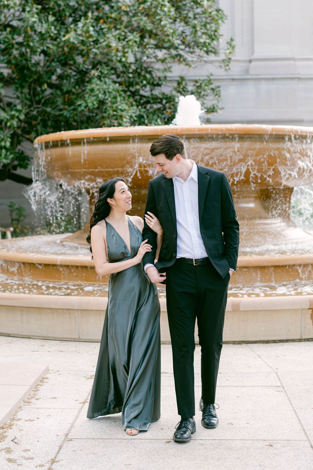 A couple sharing a laugh while walking by a flowing fountain in formal attire.