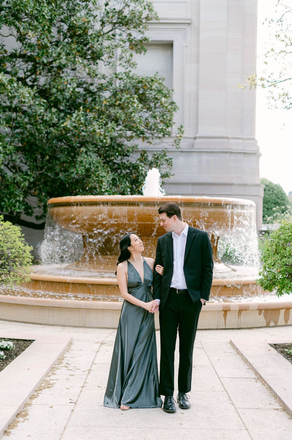 Elegant couple poses in formal attire by a fountain in a garden setting.