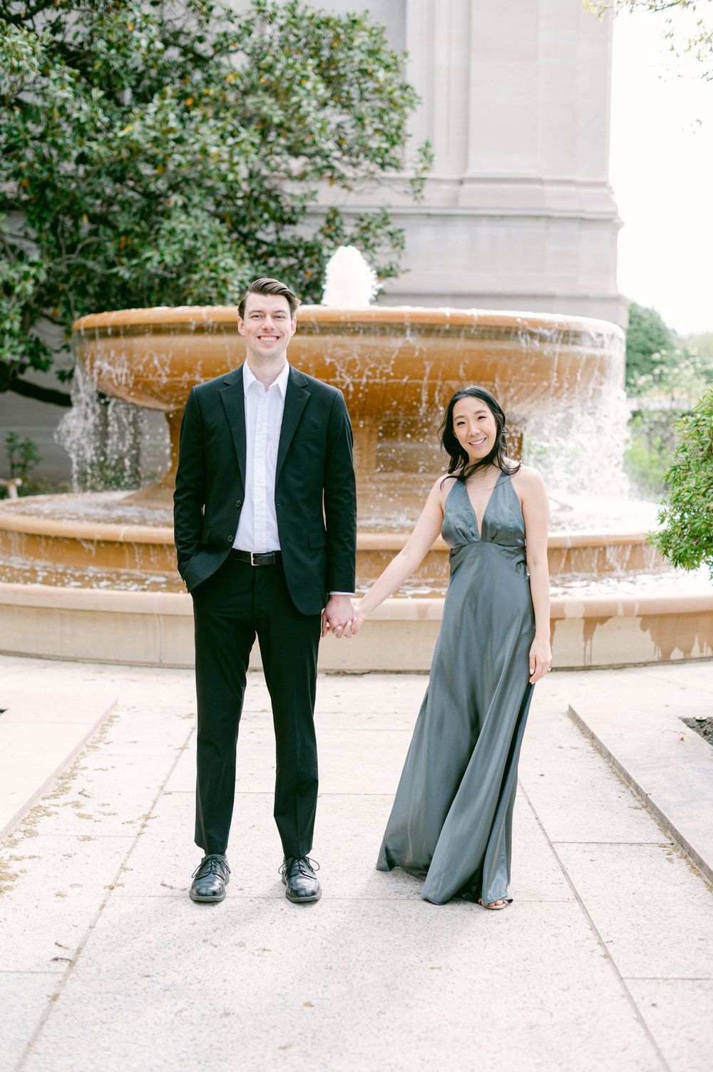 Couple in formal wear holds hands while standing in front of an ornate fountain.