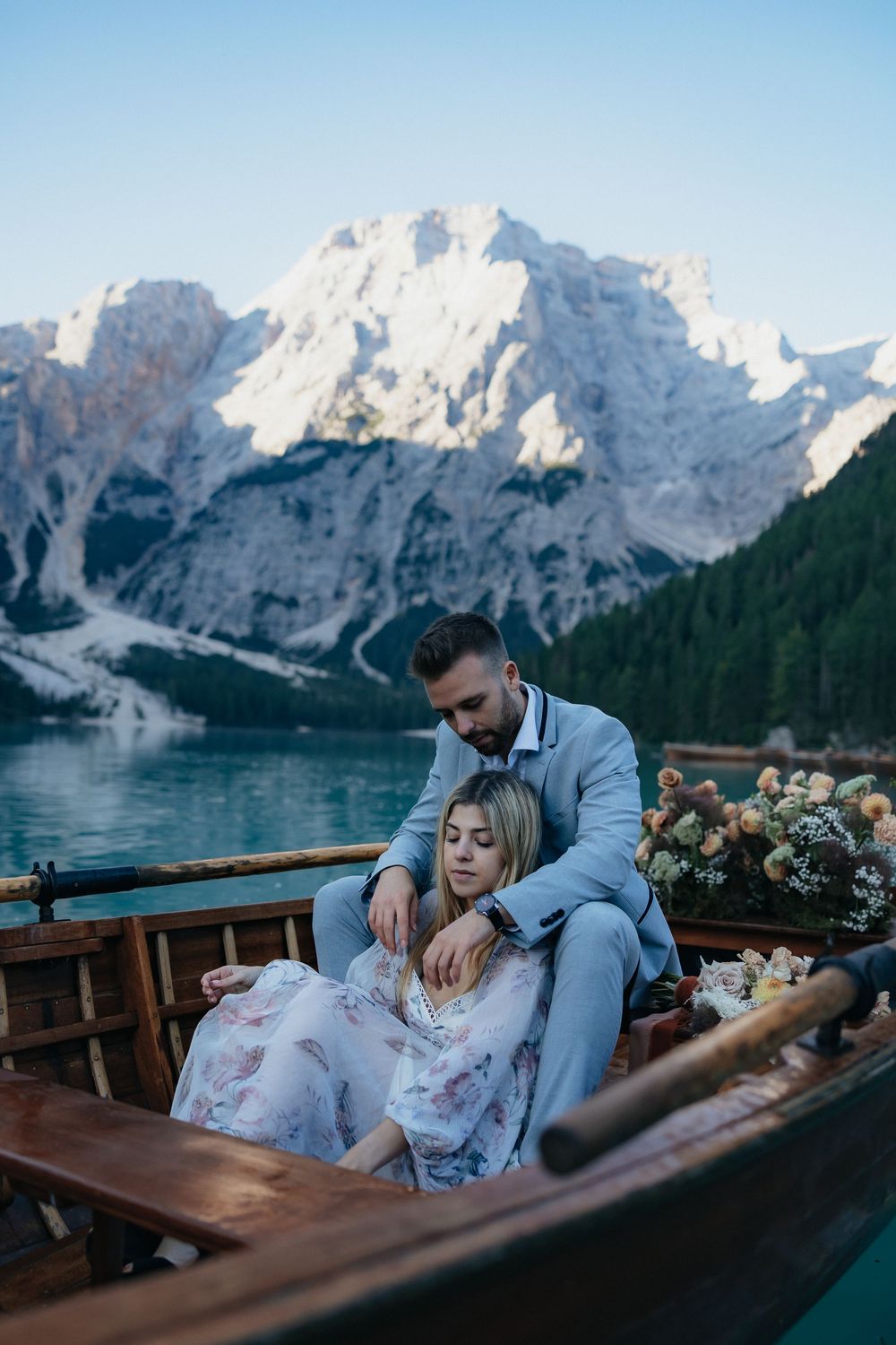 Couple sharing romantic moment in wooden boat on alpine lake with snow-capped Dolomites mountains in background.