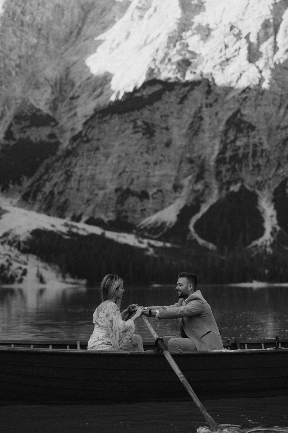 Two people sit in a wooden boat on a lake surrounded by towering mountain cliffs in dramatic black and white.
