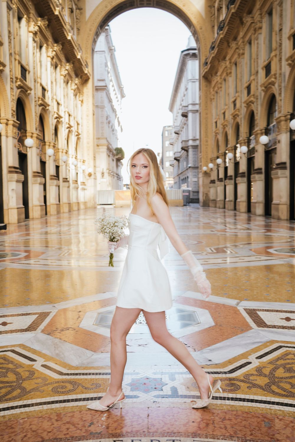 Elegant figure in white dress poses under ornate arched gallery with decorative marble floors in Milan, Italy.