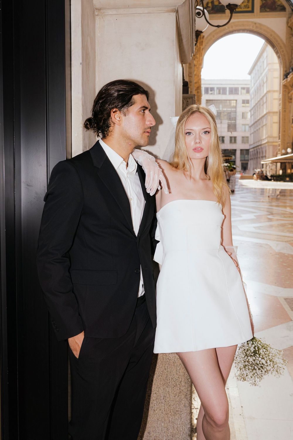 Elegantly dressed couple in black suit and white mini dress pose together in an archway on a rainy city street.