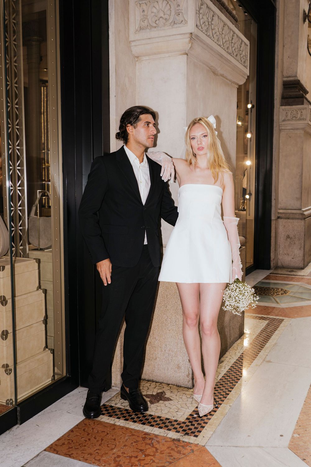 A couple stands in formal attire outside ornate doors, with the woman in a white mini dress and man in a black suit.