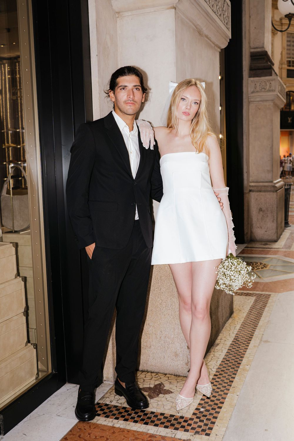 A couple in formal attire stands together in an elegant hallway with checkered floor and ornate walls.