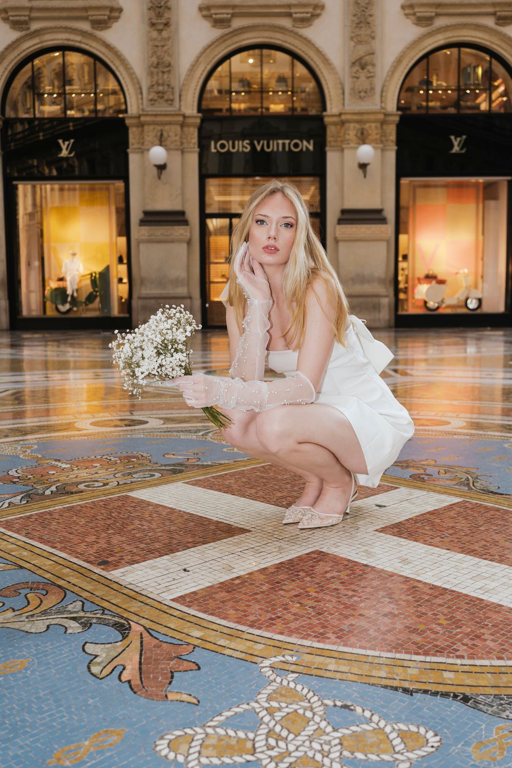 Model in white ensemble poses on ornate floor mosaic at luxury Louis Vuitton storefront at night.