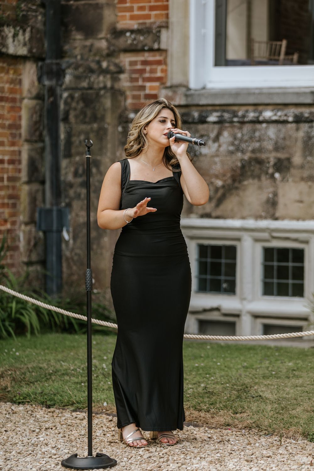 A performer in a black formal dress sings into a microphone while standing outdoors near a brick building.