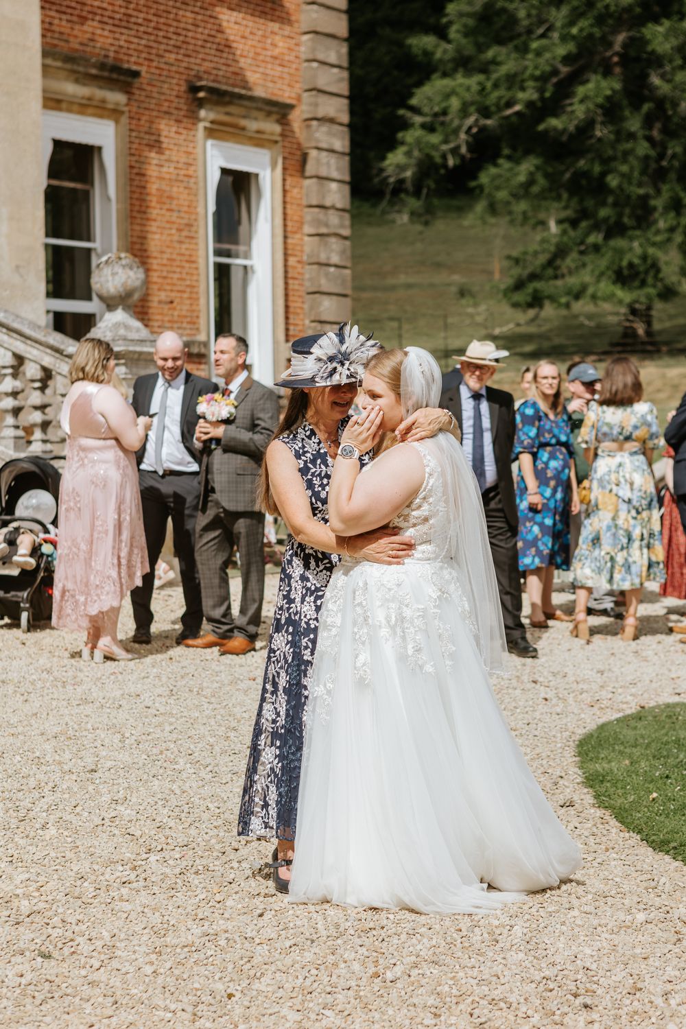 Wedding guests gather outside a brick manor house as a bride in white embraces someone during an outdoor celebration.