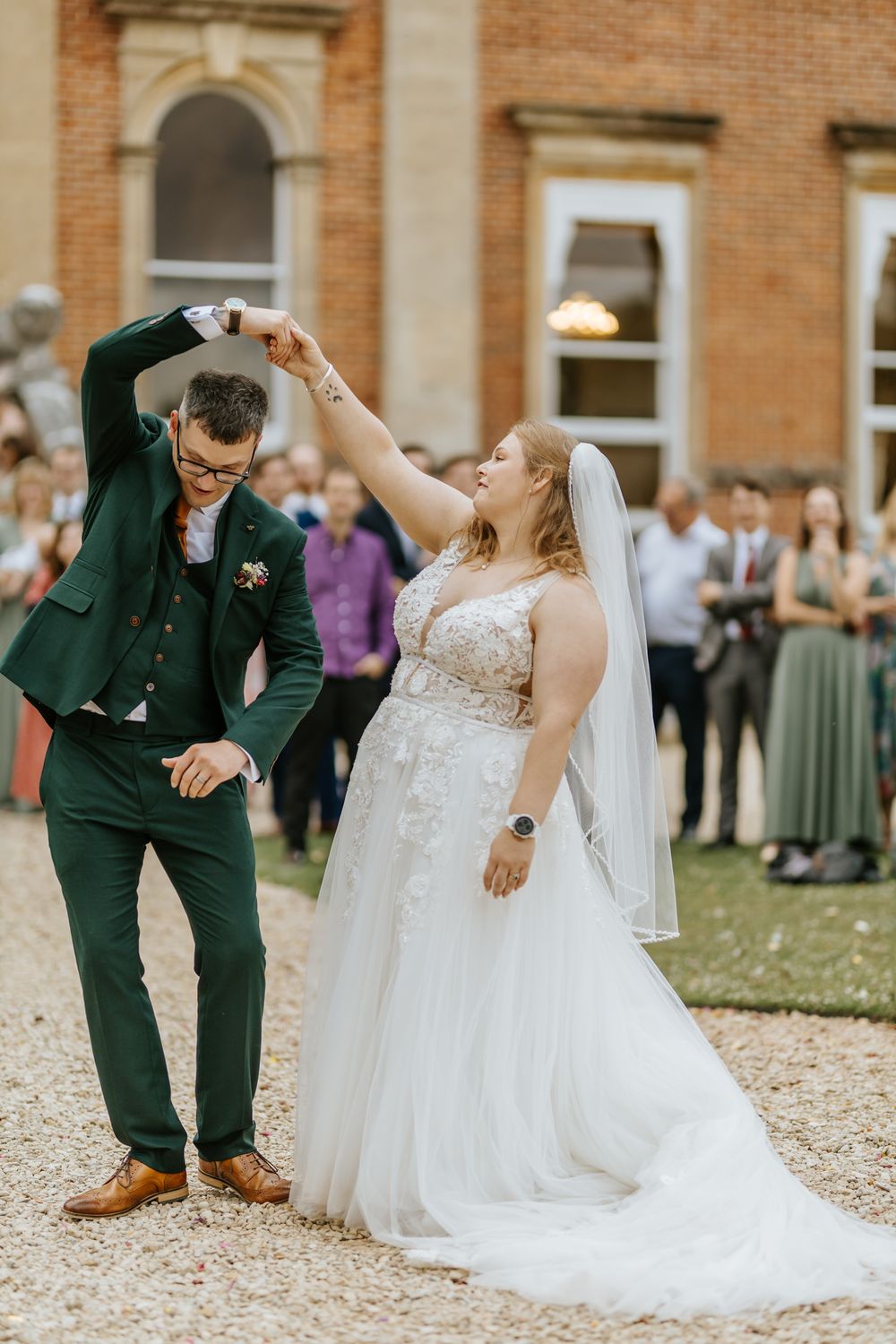Couple in wedding attire smiles and dances playfully outdoors in front of a brick building.