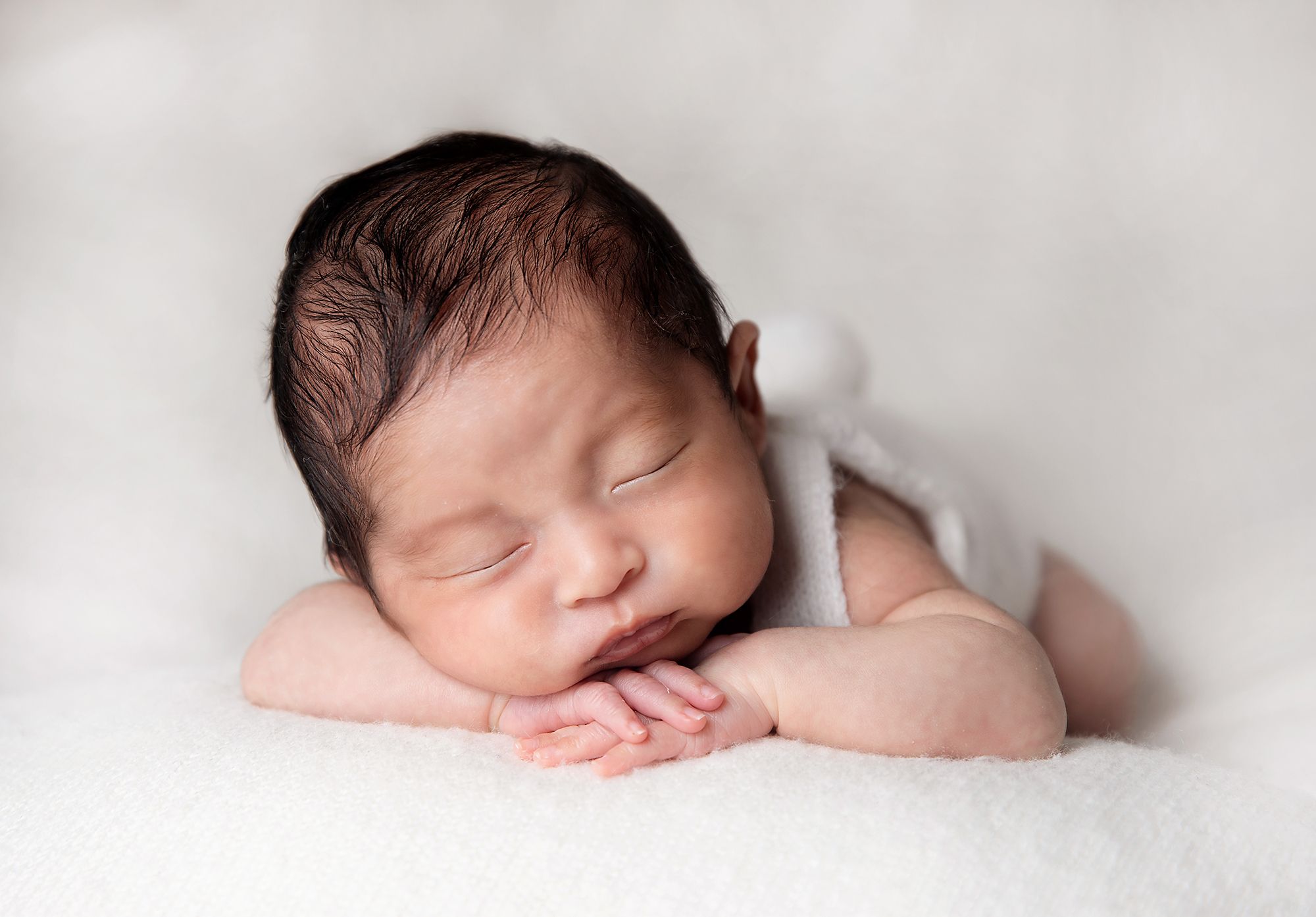 A peacefully sleeping newborn baby with dark hair rests against a soft white blanket. During a newborn photoshoot in Bexley