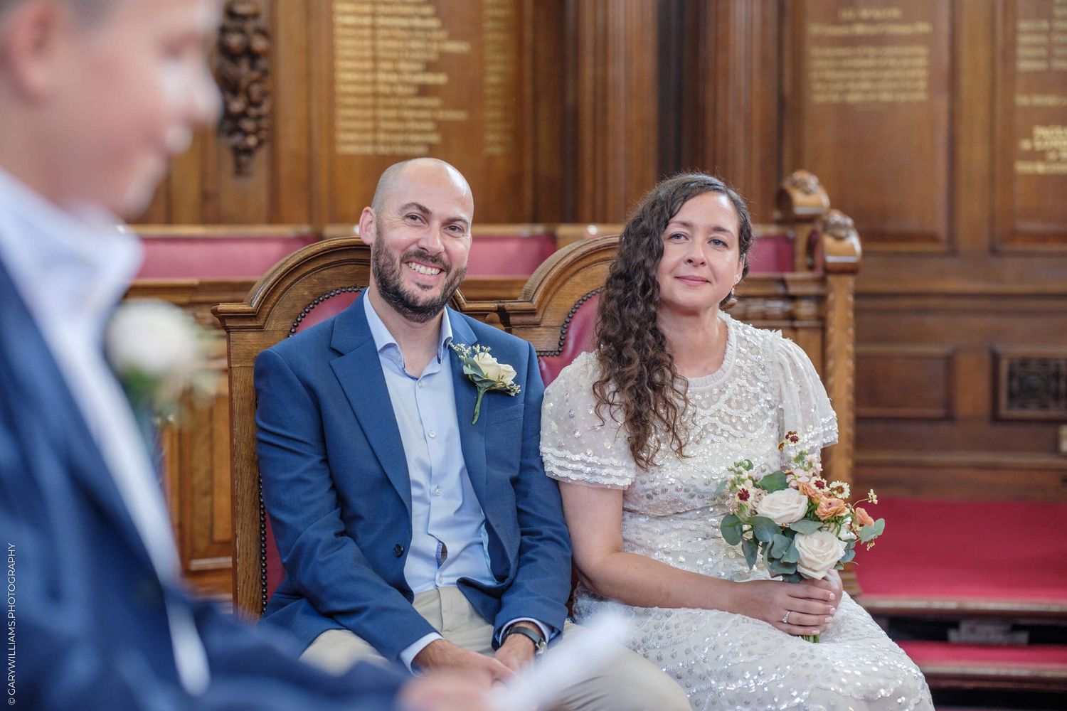 A couple sits together during an intimate wedding ceremony in a traditional wood-paneled venue with a red carpet aisle.