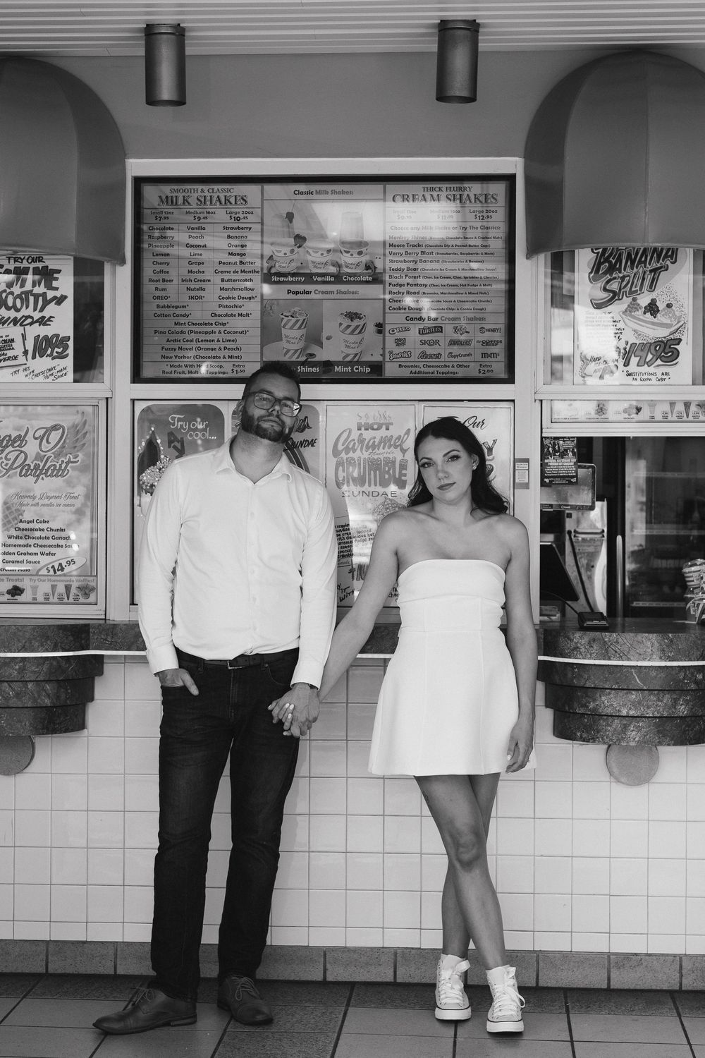 Black and white photo of a couple standing at an ice cream shop counter with vintage menu boards and signs on the wall.