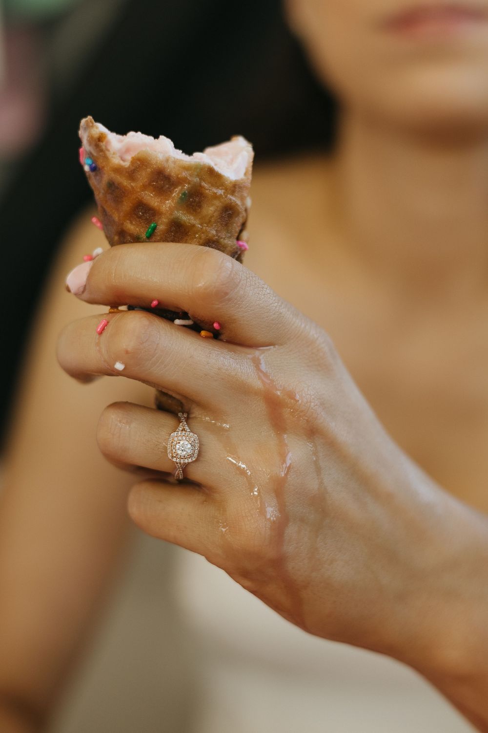 A hand wearing a diamond engagement ring holds a dripping ice cream cone with sprinkles.