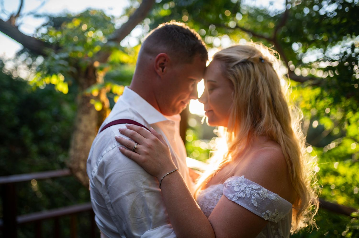 A romantic couple embraces in golden sunlight surrounded by lush green trees as the sun sets behind them.