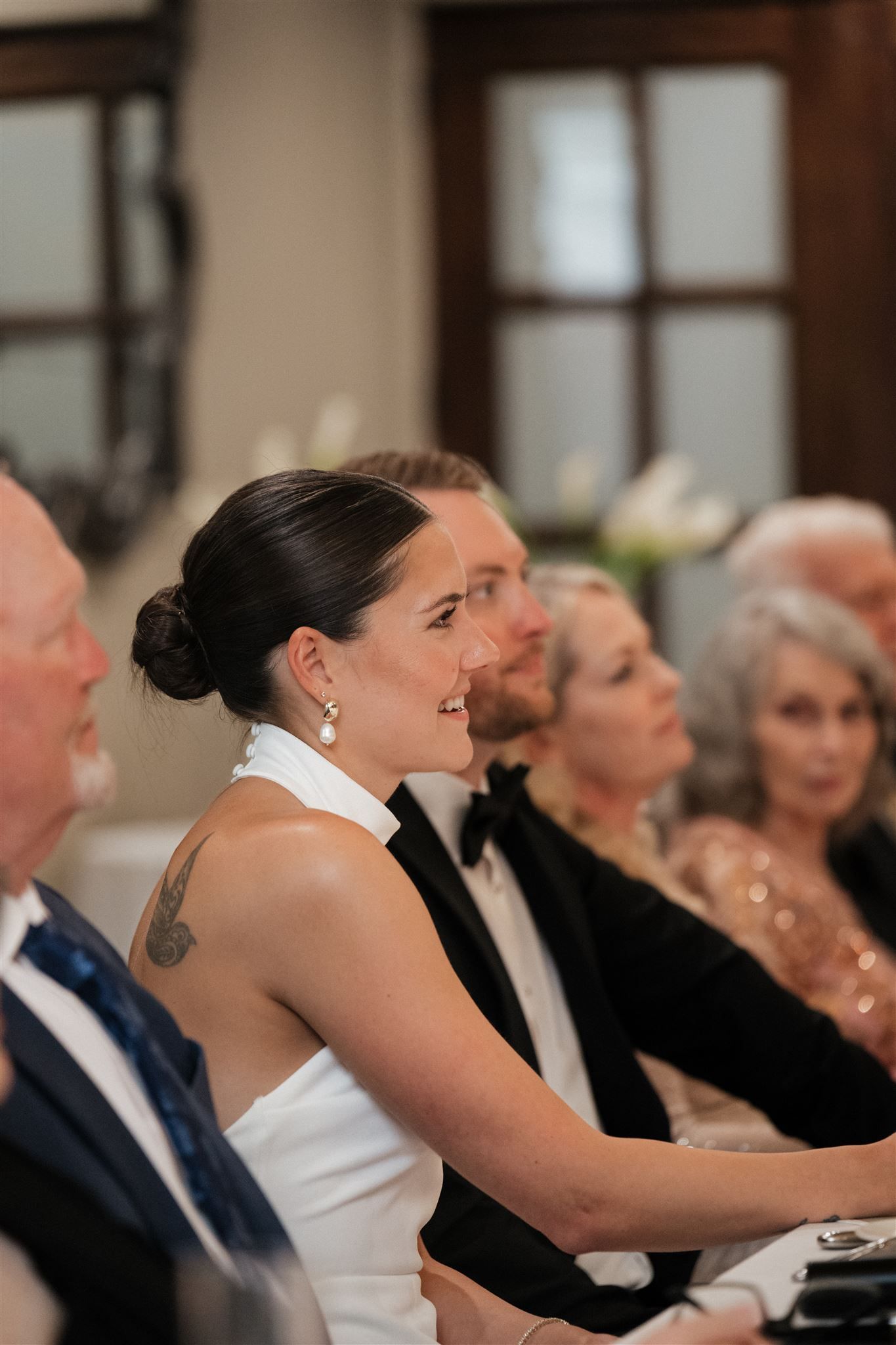 Wedding guests laugh and smile while listening to speeches during a reception celebration.