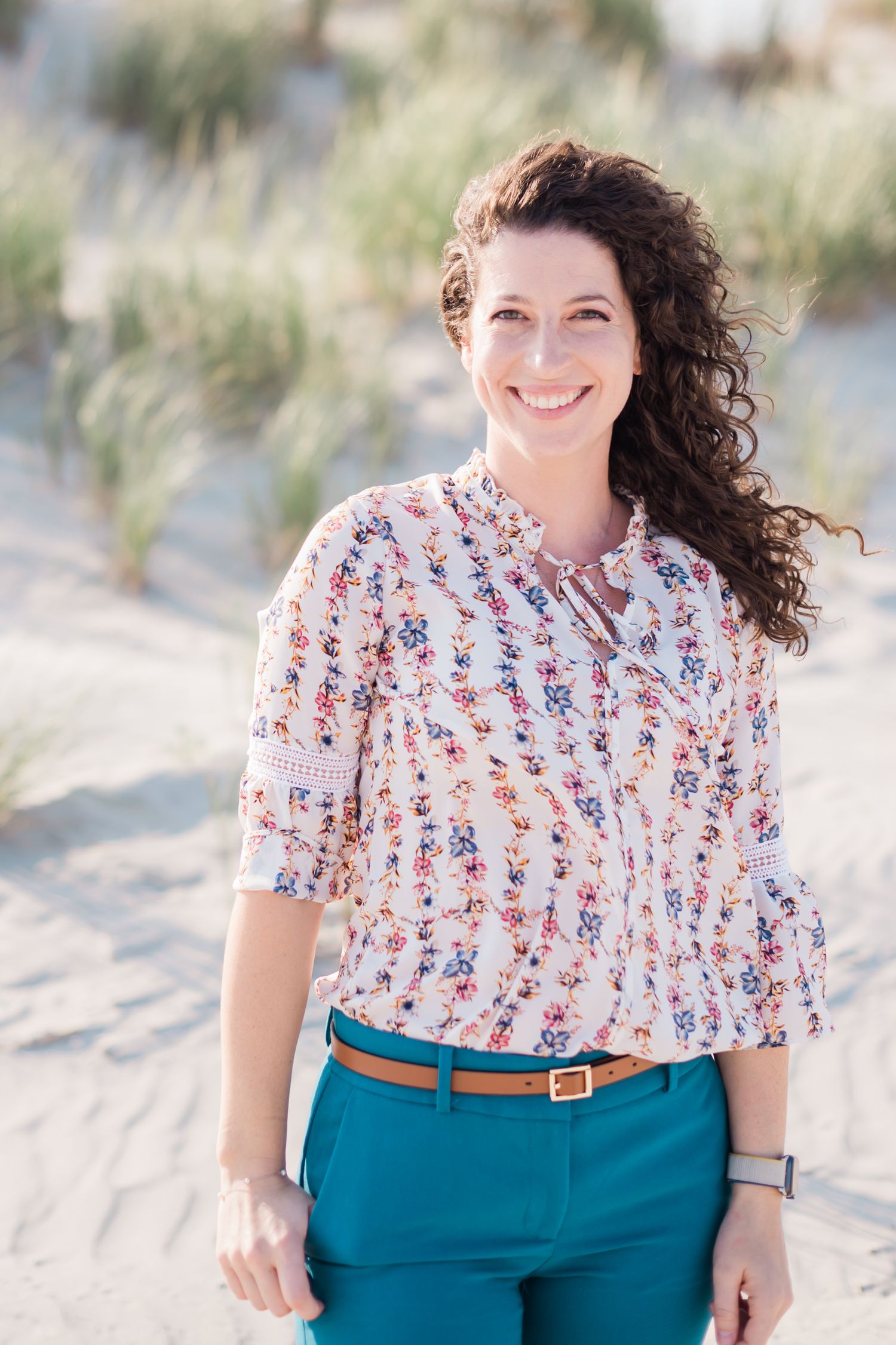 Person wearing a white floral print top and teal pants poses outdoors at the beach in a series of casual portraits.