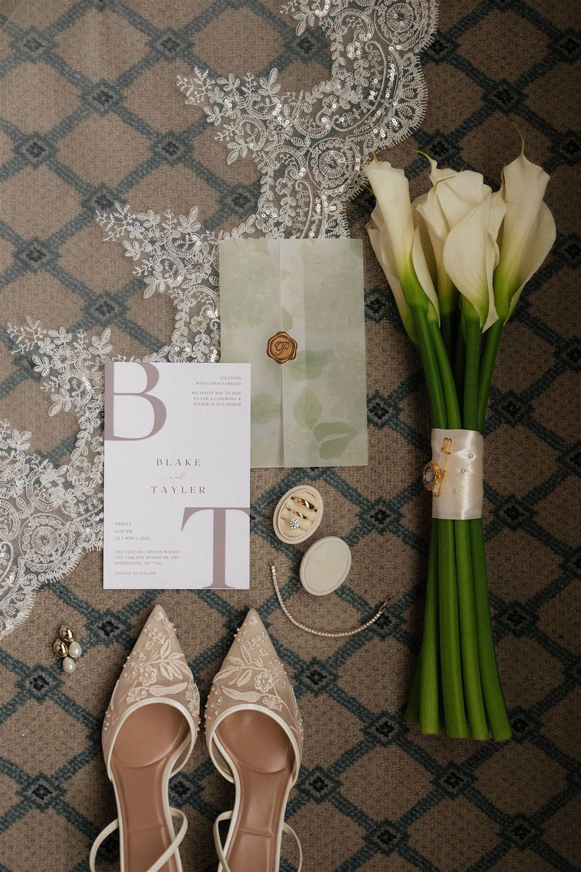 Wedding details flatlay with white calla lilies, lace veil, beige pointed shoes, and elegant invitations on patterned carpet.