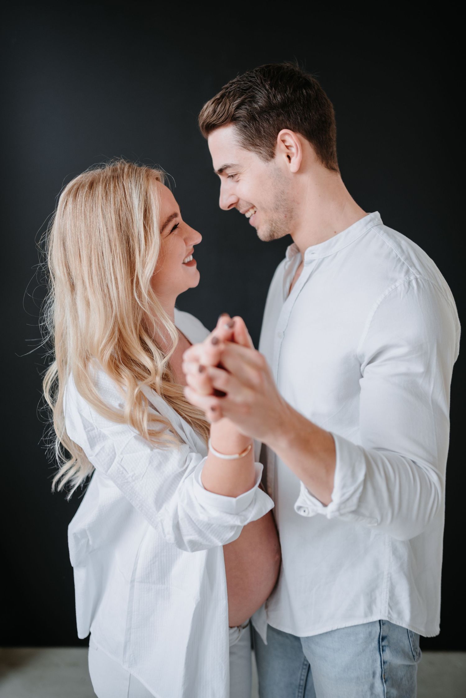 A couple wearing matching white shirts holding hands and smiling at each other against a dark background.