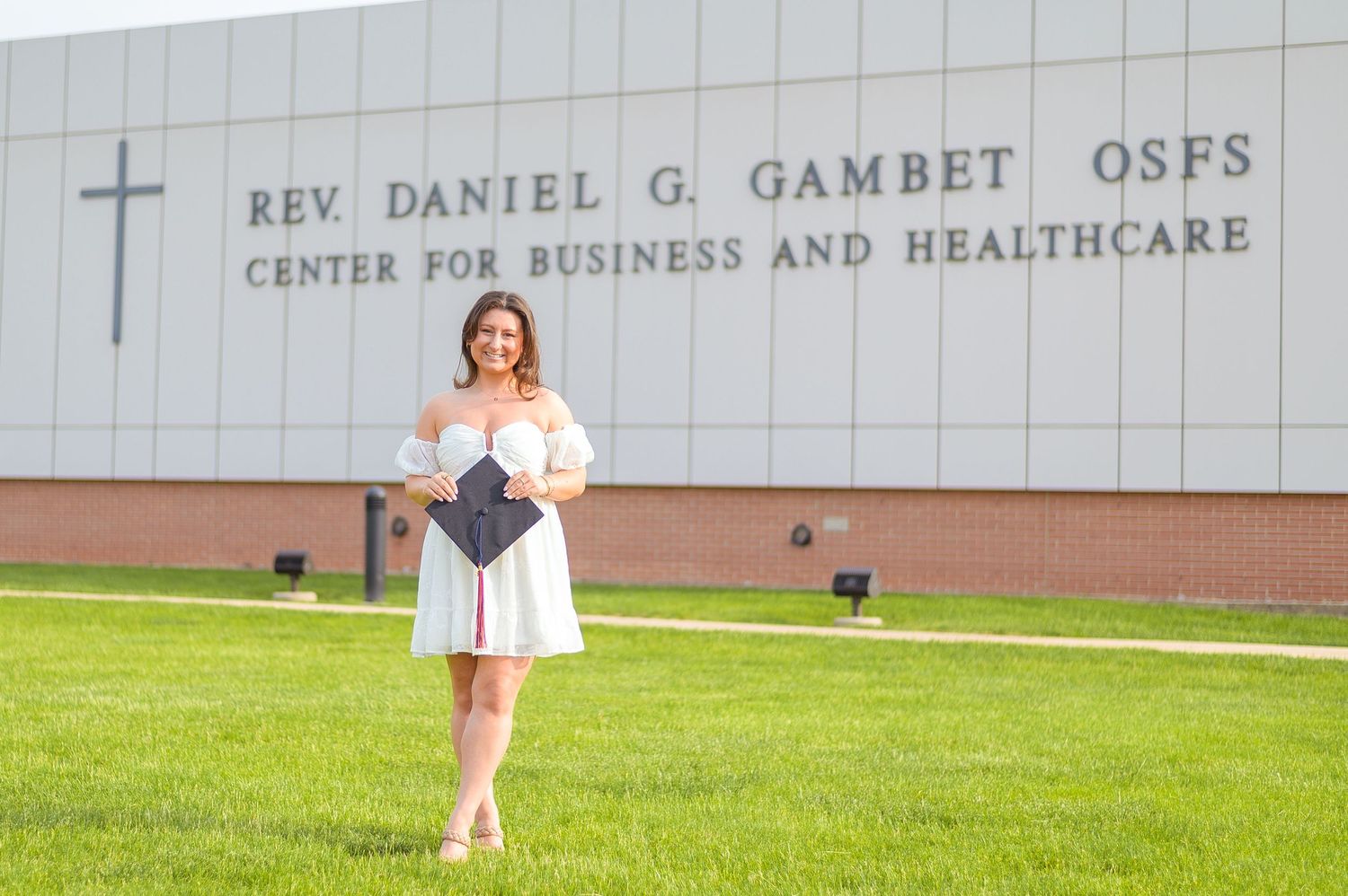 Graduate in white dress poses with diploma outside the Rev. Daniel G. Gambet Center.