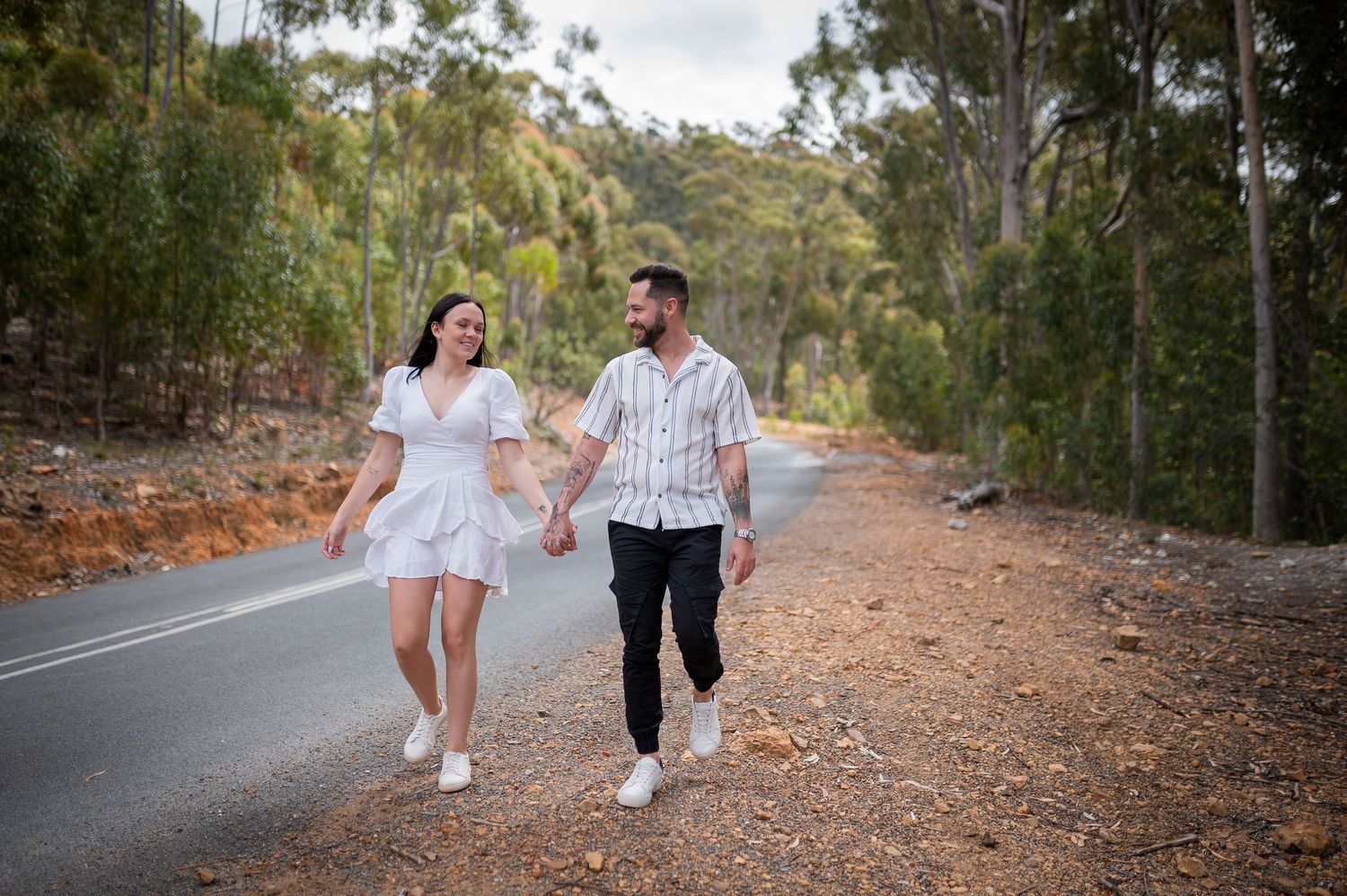 Two people walk hand in hand on a tree-lined dirt path as sunlight filters through the forest.