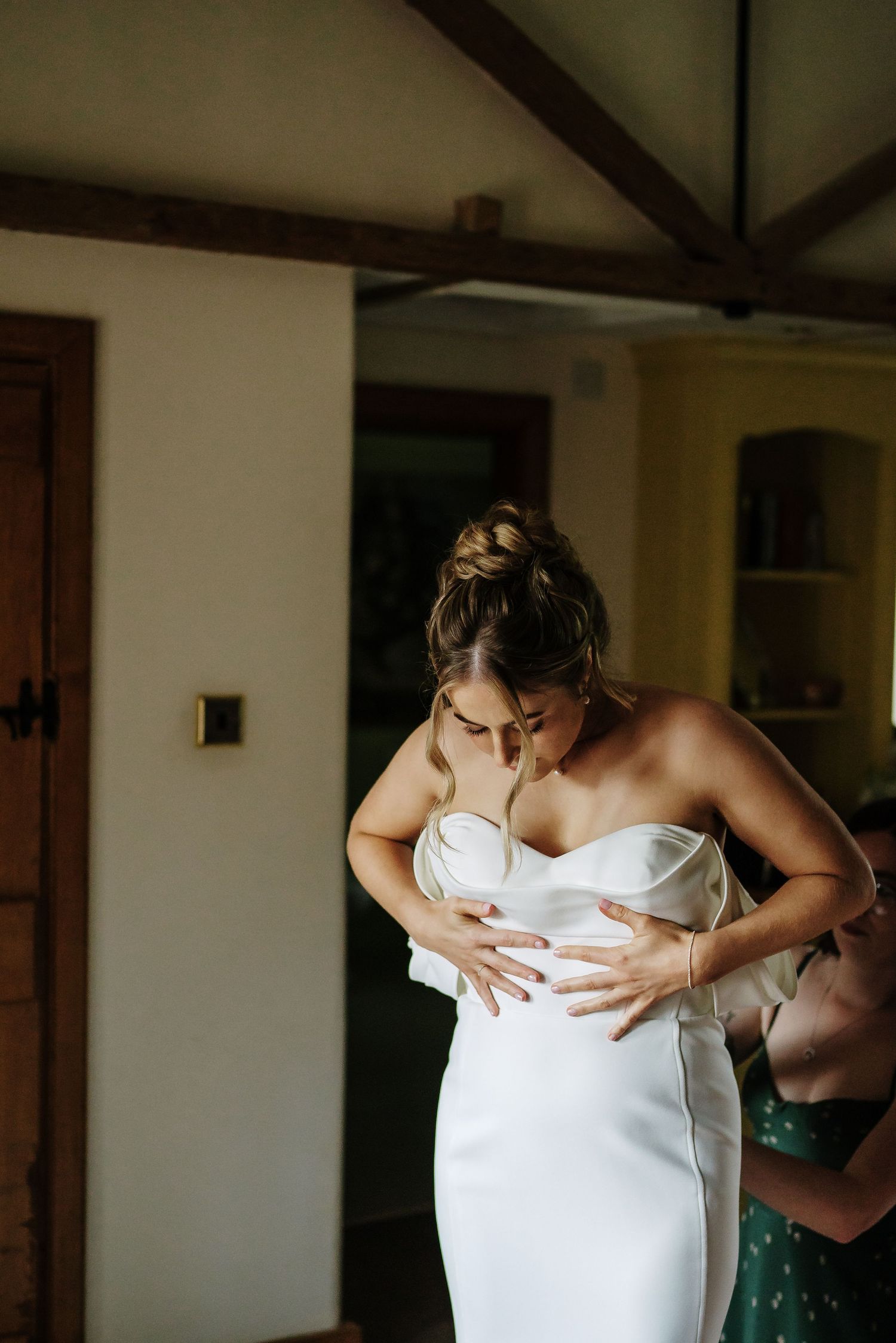 A woman in a form-fitting white strapless dress being helped into her wedding gown in a rustic room setting.