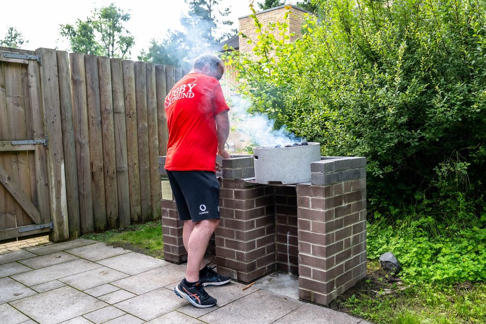 Someone wearing shorts and a red shirt tends to a brick barbecue grill with smoke rising in a backyard garden setting.