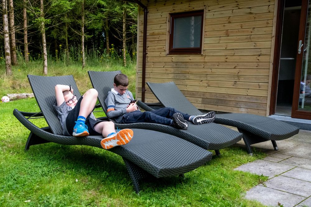 Two black lounge chairs sit on a grassy lawn outside a rustic wooden cabin on a sunny day.