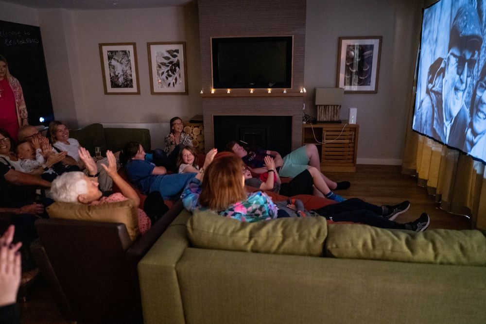 Group watching a movie together on a large sofa in a dimly lit living room with wall art and string lights.