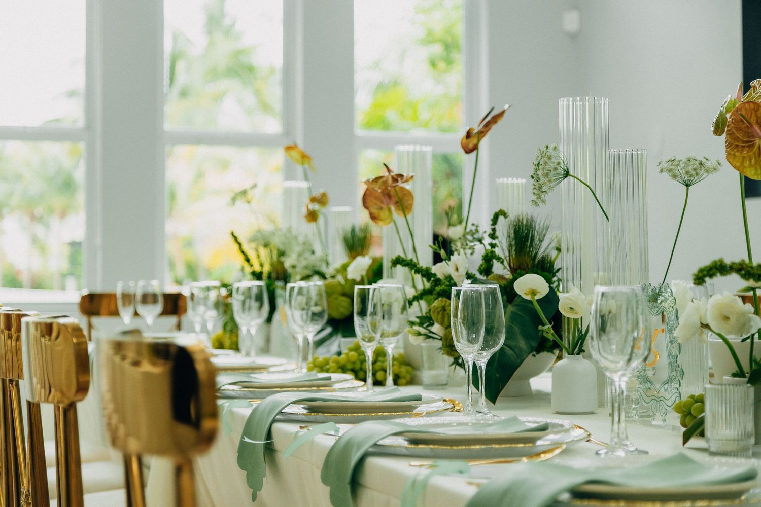 Elegant wedding reception table setting with gold chairs, white flowers, and crystal glassware in a bright room.