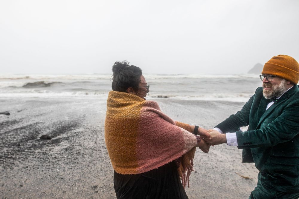 Two people hold hands on a moody winter beach with crashing waves in the background.