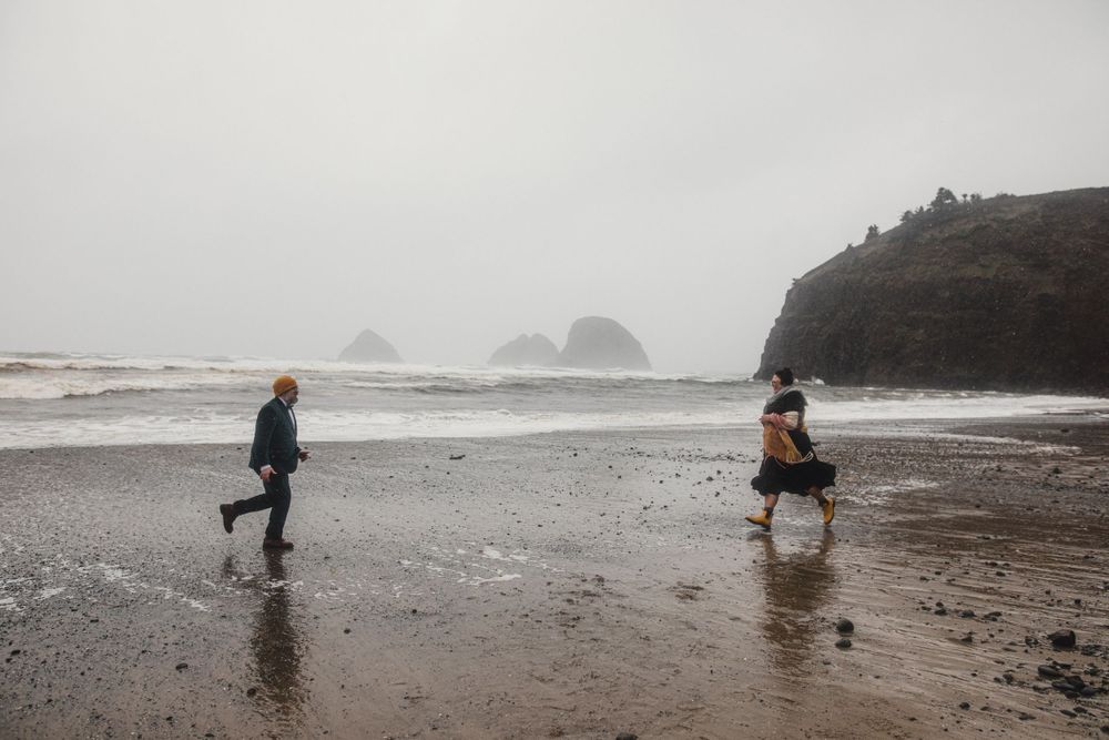 Two people run playfully along a dramatic coastline with rocky cliffs and ocean waves.