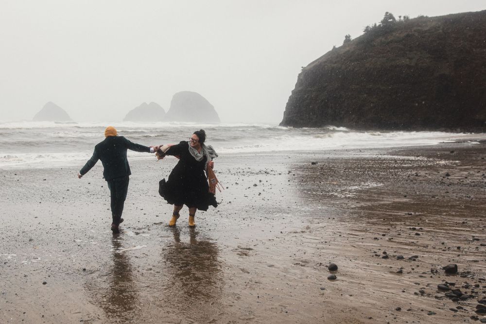 Couple dances freely on wet sand with dramatic coastal cliffs and ocean waves behind them.