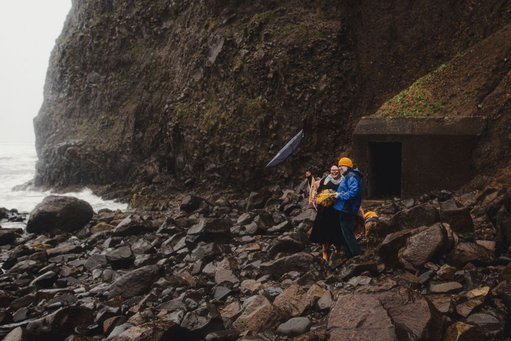 Person in bright yellow beanie sits among rocky coastal terrain near a seaside cave.