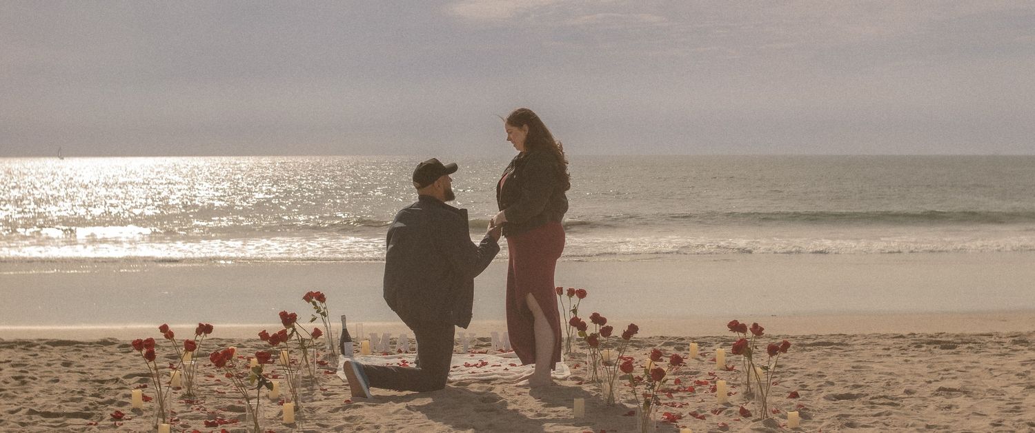 A romantic beach proposal scene at the Newport Pier with rose petals and roses with a Marry Me sign