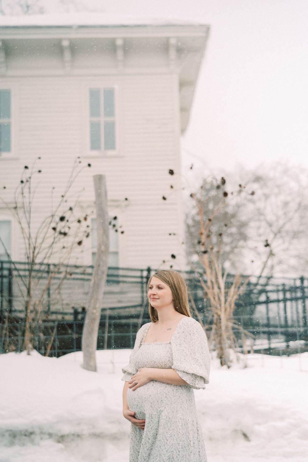 Someone in a lace maternity dress stands in the snow outside a white house with blue shutters during winter.