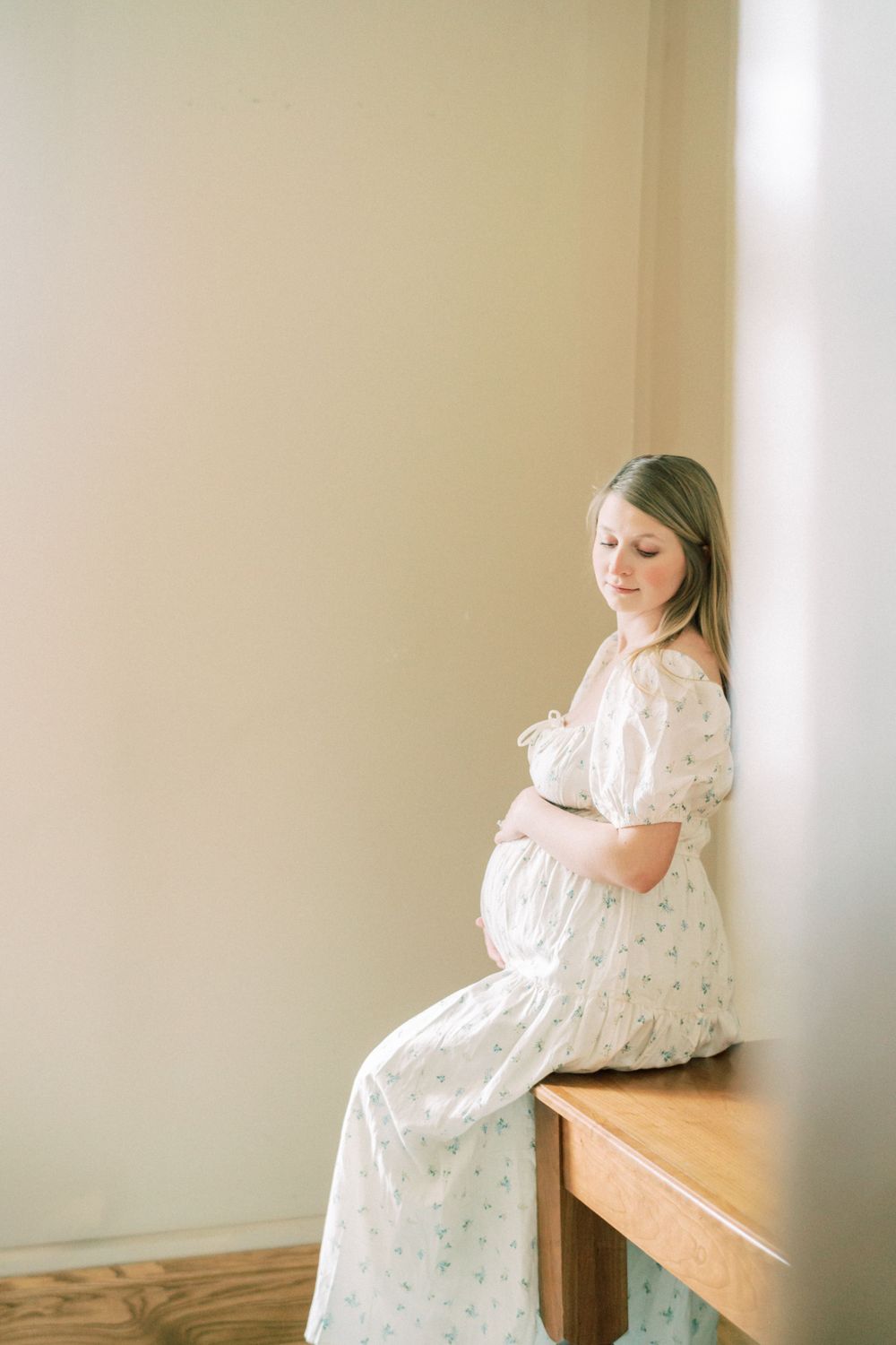 Indoor maternity portrait showing a person in a flowing white dress sitting on a wooden table in gentle lighting.