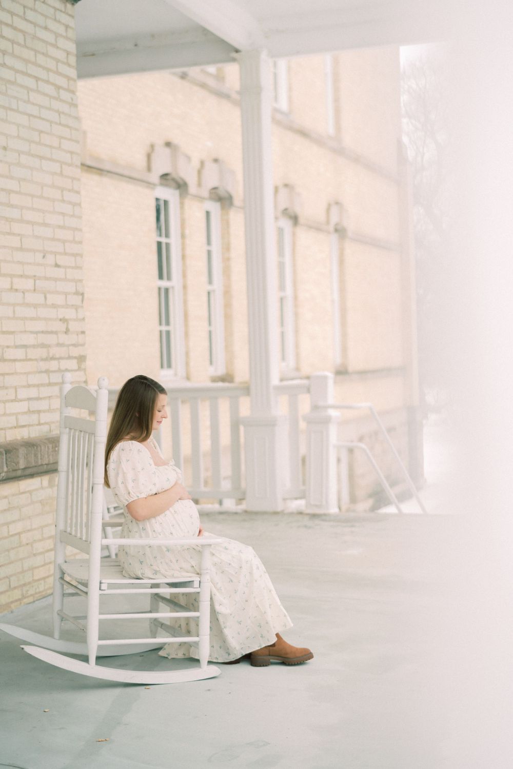 A person in a white lace dress sits peacefully on a white rocking chair on an elegant porch with architectural details.
