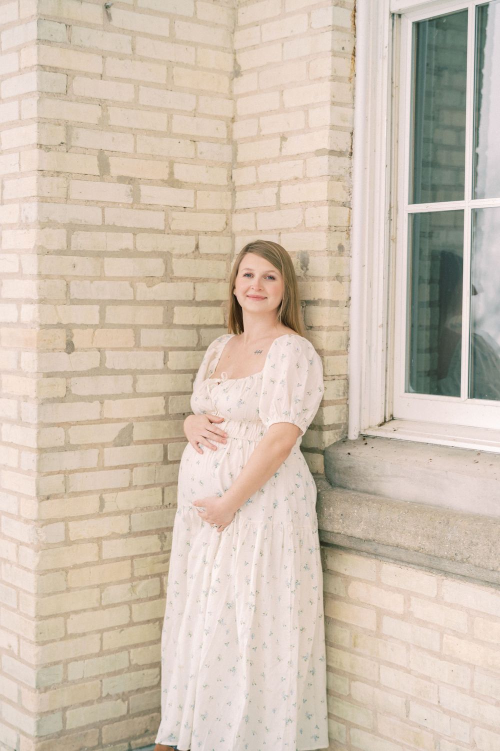 A person in a white lace maternity dress stands against a light brick wall next to a window in soft lighting.